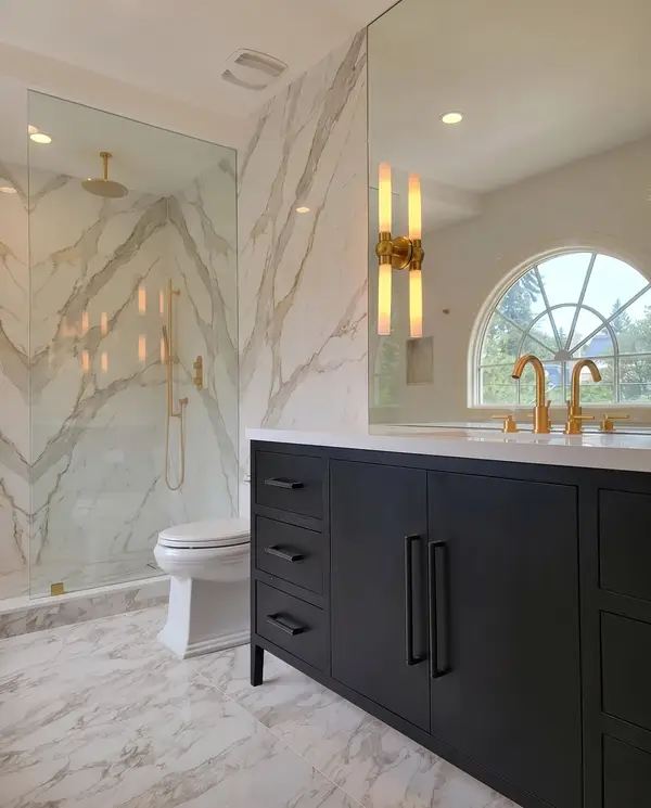 Black vanity with gold faucets under an arched window, marble-look slab shower