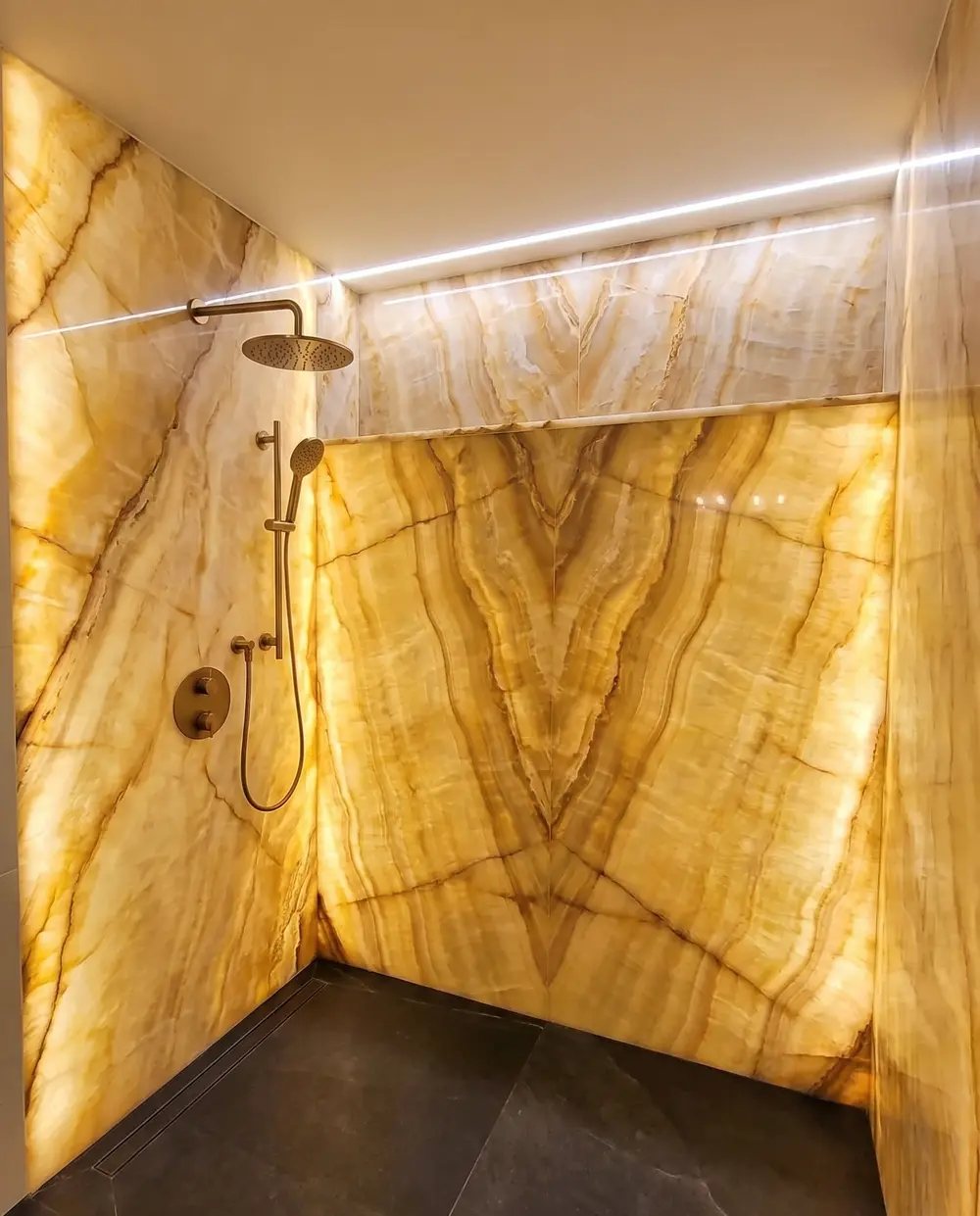 Backlit onyx-look slab shower with brushed brass fixtures and a charcoal floor